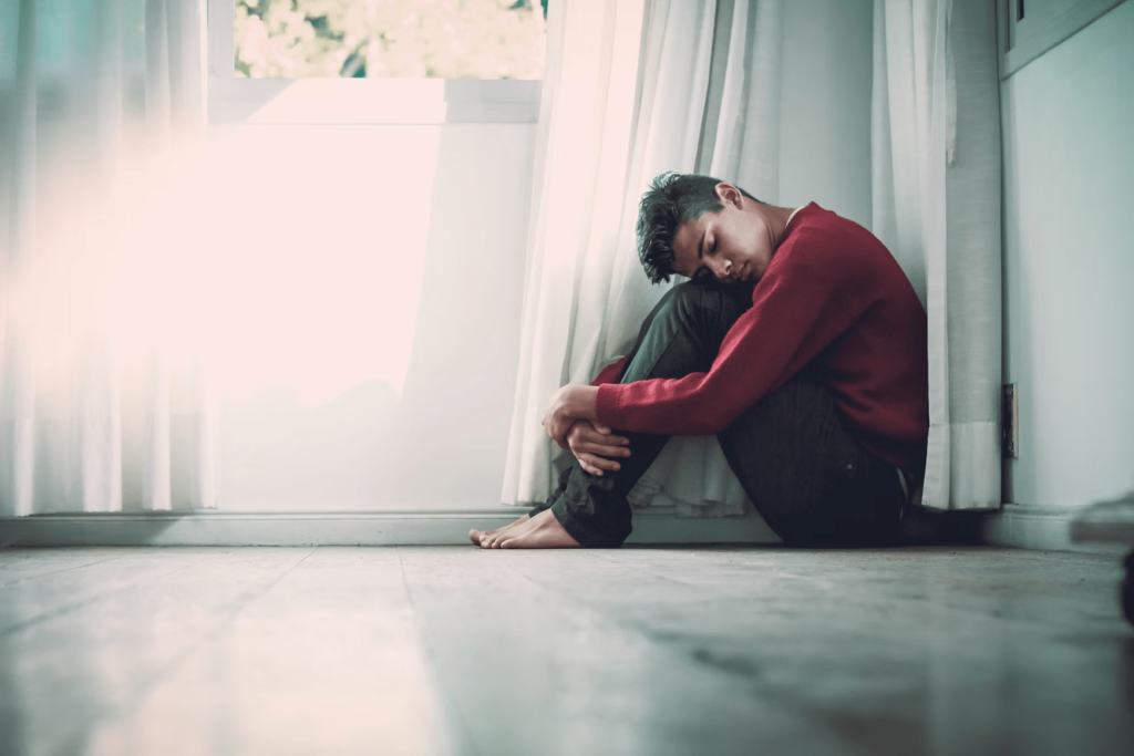 A person huddled on the floor with their arms around their legs as if in distress, perhaps in need of mental health care through their Magellan Mental Health Rehab Treatment Plan